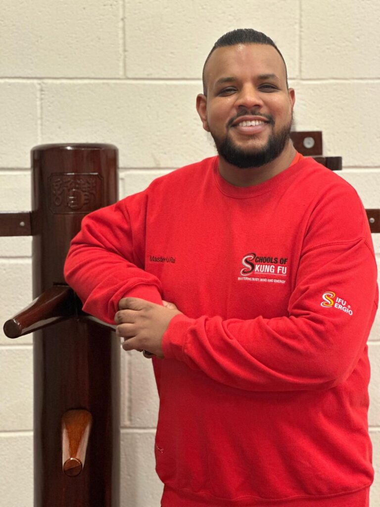 Profile picture of SiFu Kul wearing a red master sweatshirt, leaning against a Wing Chun wooden dummy, smiling with a relaxed and confident expression.