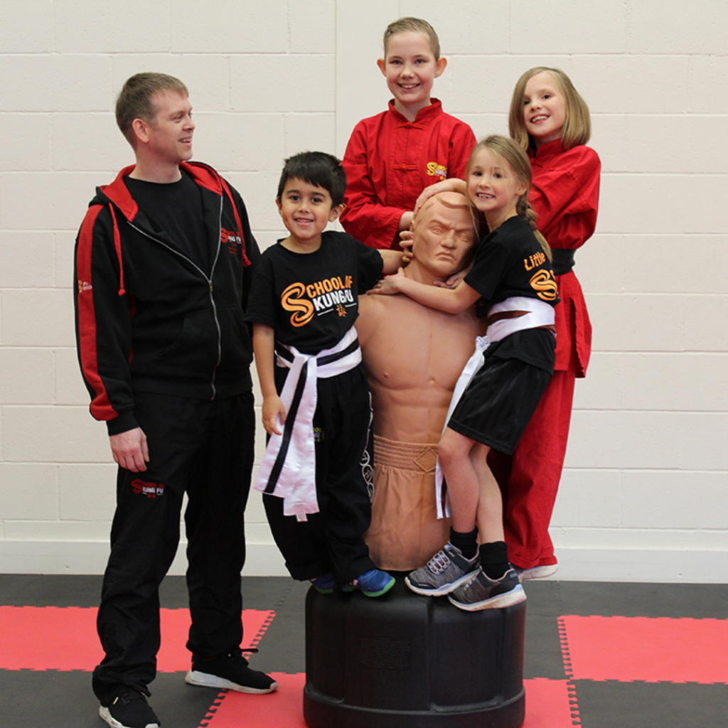 four children standing on a martial arts training dummy, with instructor on the ledt